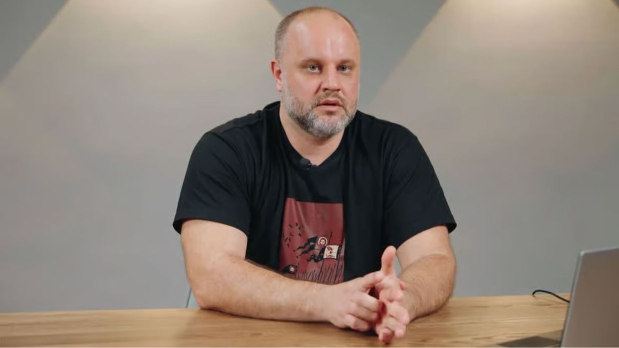 Чоловік із короткою сивою бородою в чорній футболці сидить за столом перед камерою.