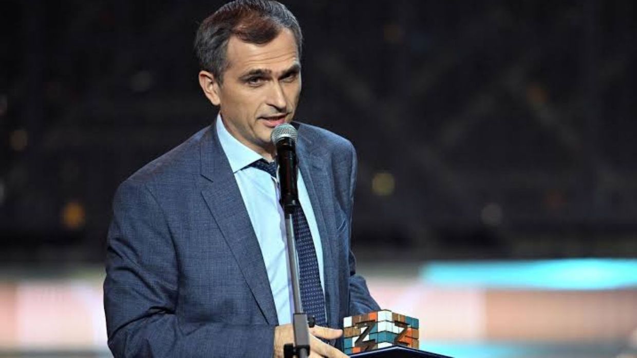 Чоловік у синьому костюмі виступає перед мікрофоном і тримає предмет із літерами Z.