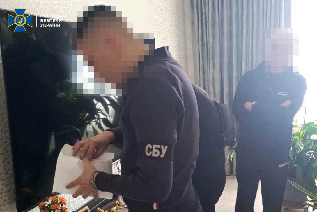 Чоловік із пов’язкою «СБУ» переглядає документи біля шафи, позаду стоїть інший чоловік.
