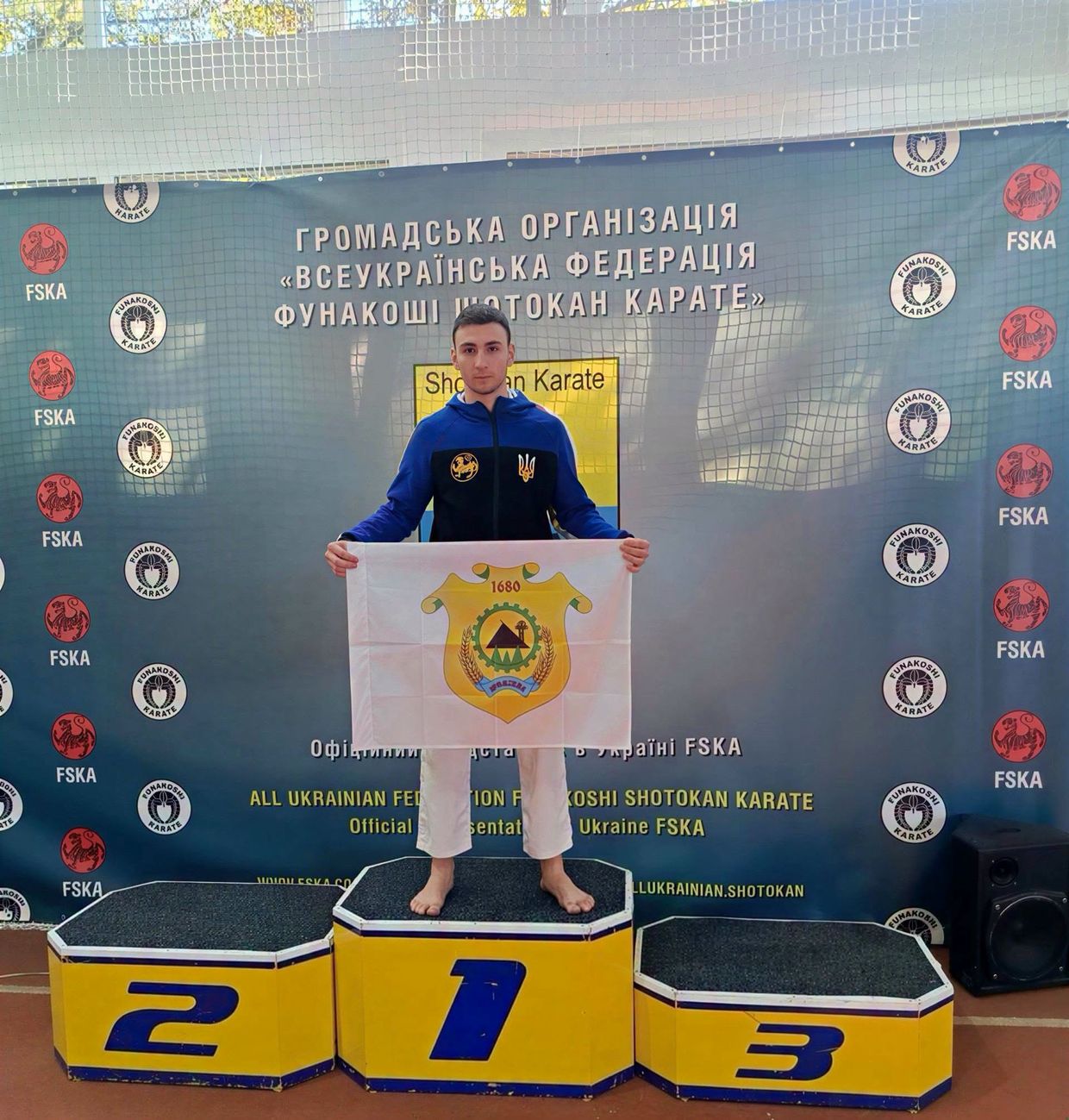 Хлопець у спортивній формі стоїть на першій сходинці п’єдесталу, тримаючи прапор із гербом Кремінної.