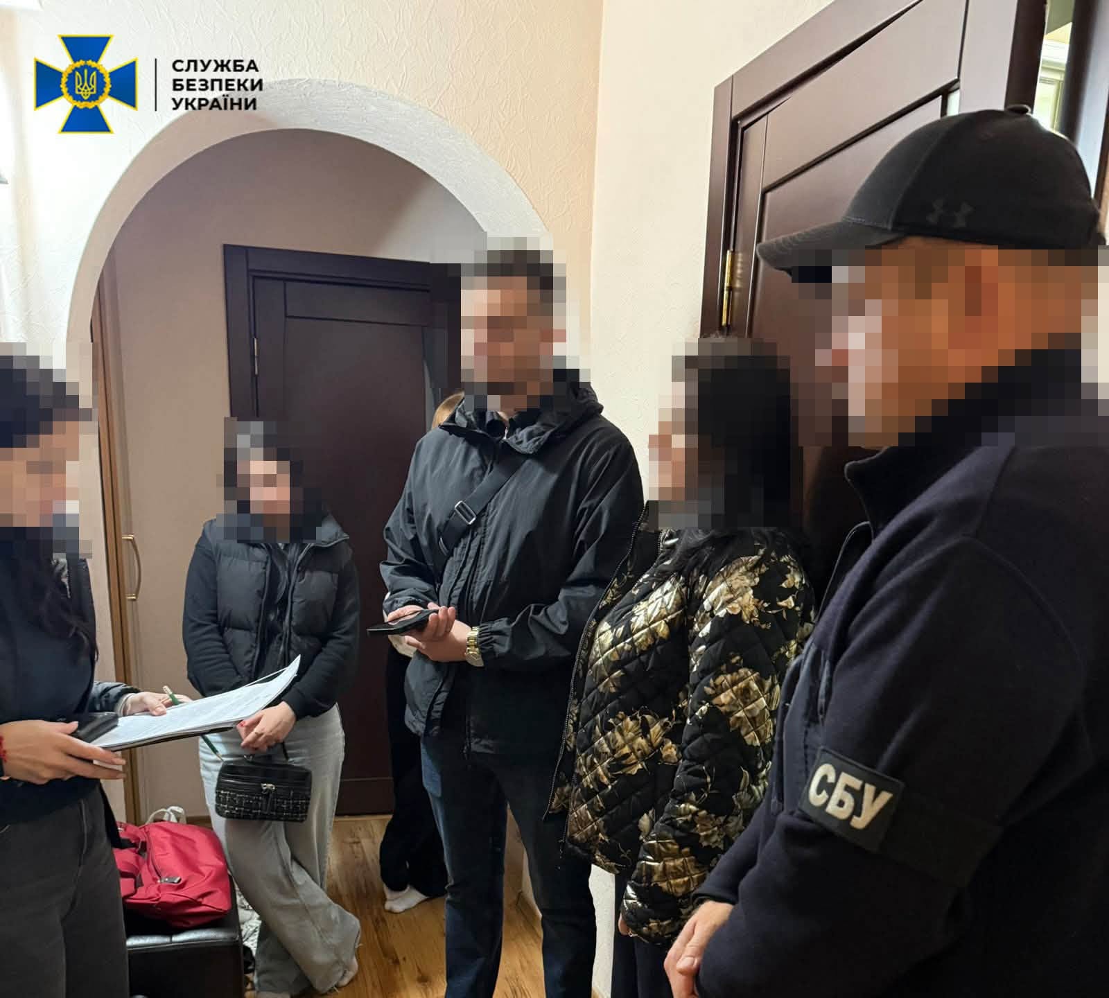 У приміщенні стоять кілька людей із заблюреними обличчями, одна з них тримає документи.