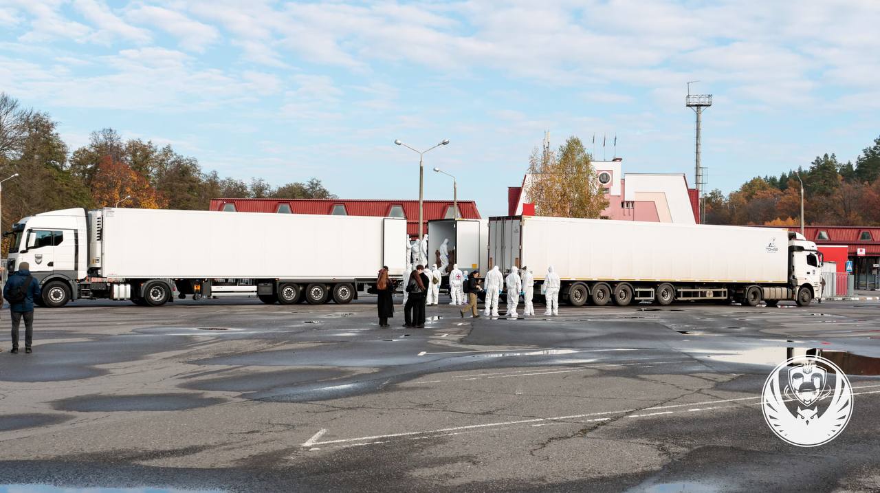 Дві великі білі вантажівки та група людей у захисних костюмах на асфальтованому майданчику.